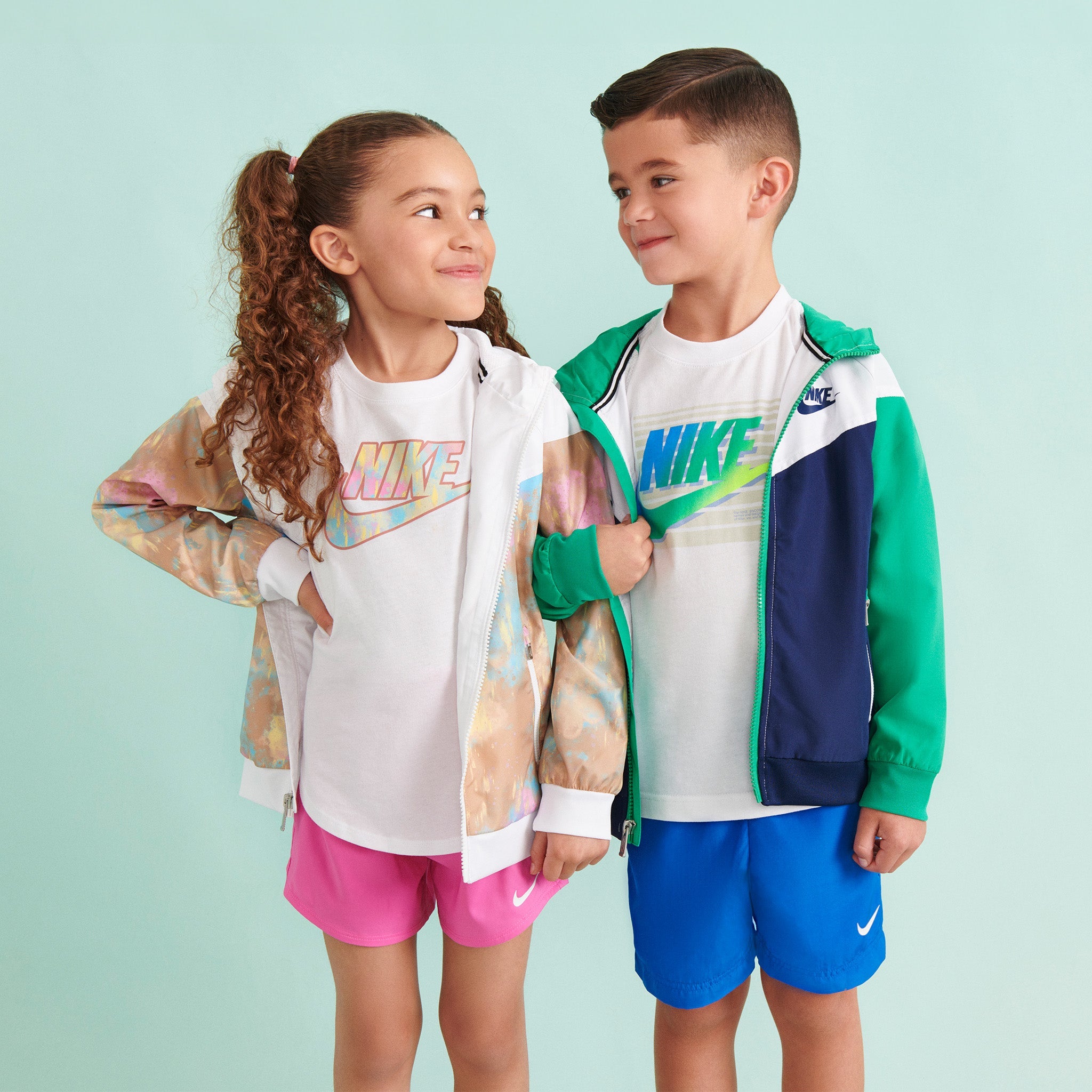 two kids smiling at each other wearing white Nike t-shirts, multicolored jackets and colorful Nike shorts